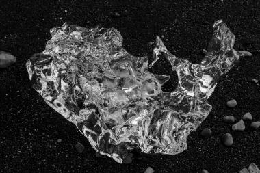Close-up of a large, jagged, transparent crystal or ice shard resting on coarse black sand with small pebbles, showing bright reflective surfaces and deep shadows.