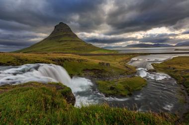 Landscape of Kirkjufell mountain in Iceland with a cascading waterfall in the foreground, a grassy riverbank, and dark, stormy clouds overhead.