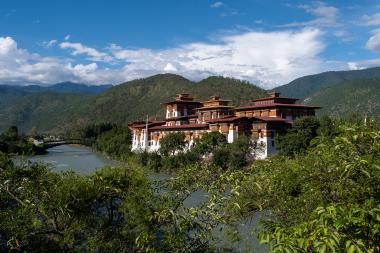 Wide landscape view of Punakha Dzong, a large white-and-red Bhutanese fortress monastery with tiered golden roofs, perched beside the Mo Chhu River; green shrubs fill the foreground, a bridge crosses the river to the left, and forested mountains rise under a blue sky with scattered clouds in Punakha, Bhutan.