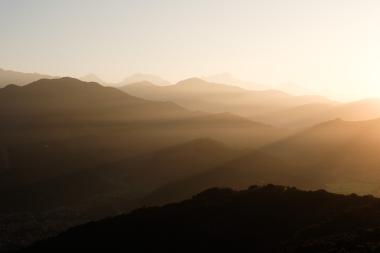 A sweeping view of distant mountain ranges at sunset, with soft golden light casting long shadows across multiple ridges and a hazy sky.