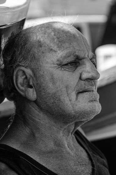 Close-up black-and-white portrait of an older man in profile. The screen-reader description: an elderly, weathered man with a closely trimmed stubble and receding hairline looks to the right with a thoughtful expression. Wrinkles and textured skin are visible on his forehead, around his eyes and along his neck. The background is out of focus and suggests an outdoor setting near boats or a harbor.