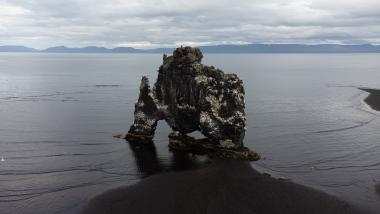 A tall, jagged basalt rock formation forming a natural arch at the edge of a black sand beach, with a calm sea and distant mountain range under gray clouds; a small seagull is visible near the left edge.