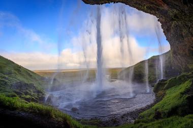 Behind the Seljalandsfoss waterfall in Iceland, water streams down from the cliff inside a mossy cave, creating a veil of mist over a shallow pool. The landscape beyond shows green hills, a wide plain, and a bright blue sky with soft clouds.