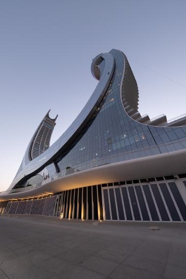 A futuristic glass skyscraper with sweeping curved lines and a circular top, set against the evening sky. The lower level shows a row of tall, dark glass doors with warm ground lights, forming a modern architectural promenade.