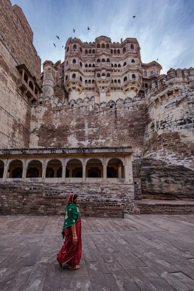 Low-angle view inside Mehrangarh Fort in Jodhpur, India: a woman in a red sari and green shawl walks across a broad stone courtyard while birds circle above the massive sandstone fort walls and carved palace façade.