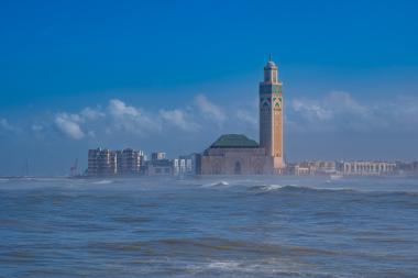 A coastal view of the Hassan II Mosque in Casablanca, Morocco. The tall minaret with green-tiled accents stands beside a large mosque with an arched entrance, set along the Atlantic Ocean. Waves crash in the foreground and a cityscape with modern buildings lines the coast under a bright blue sky.