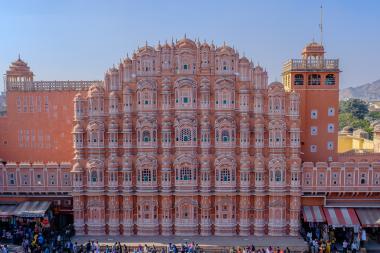 Wide, front-facing view of Hawa Mahal in Jaipur, India—an elaborate pink sandstone palace facade with many small arched windows and domed kiosks—while tourists and market stalls line the street below under a clear blue sky.