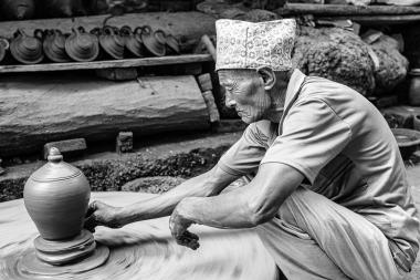Screen-reader description: An elderly man wearing a patterned Nepali hat sits at a potter's wheel, shaping a clay pot with his hands while spinning. The background shows shelves with pots and rough workshop materials.
