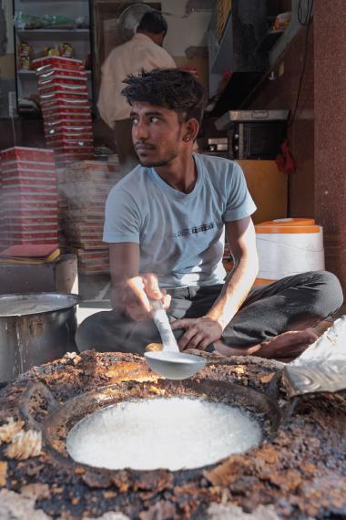 Inside a small street food shop in Bikaner, Rajasthan, a young man sits cross‑legged and uses a metal ladle to stir a large, steaming cauldron of simmering milk or sugar syrup set into a stove-like counter, while another worker moves in the background among stacked red-and-white product boxes.