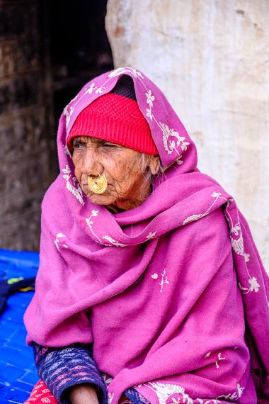 Portrait of an elderly woman seated and looking to the left, bundled in a vivid pink embroidered shawl over a red knit cap, with a large gold nose ring; the background is a rough stone wall and a blue fabric mat, photographed near Jodhpur, Rajasthan, India (Dec 21, 2023).