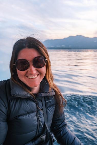 Portrait of a smiling woman in round sunglasses and a dark puffer jacket sitting on a small boat. Behind her, rippling lake water leads to a low mountain ridge under a pastel sunset sky near Udaipur, Rajasthan, India.