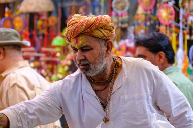 Close-up in a busy street market in Pushkar, Rajasthan: an older man wearing a patterned turban, white shirt, and bead necklaces, with a saffron tilak on his forehead, leans forward and reaches out as bright hanging handicrafts blur in the background.