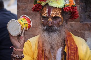 Close-up of a bearded man wearing a vibrant floral crown, gold face paint with red markings, and a yellow robe. He holds a small, colorful drum in his right hand and wears multiple bead bracelets. A brick wall is in the background.